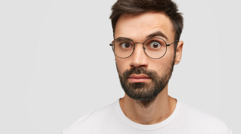 Emotive bearded young guy with thick stubble, trendy hairdo, looks with surprisement at camera, stares through glasses, dressed in casual white t shirt, expresses bewilderment and astonishment